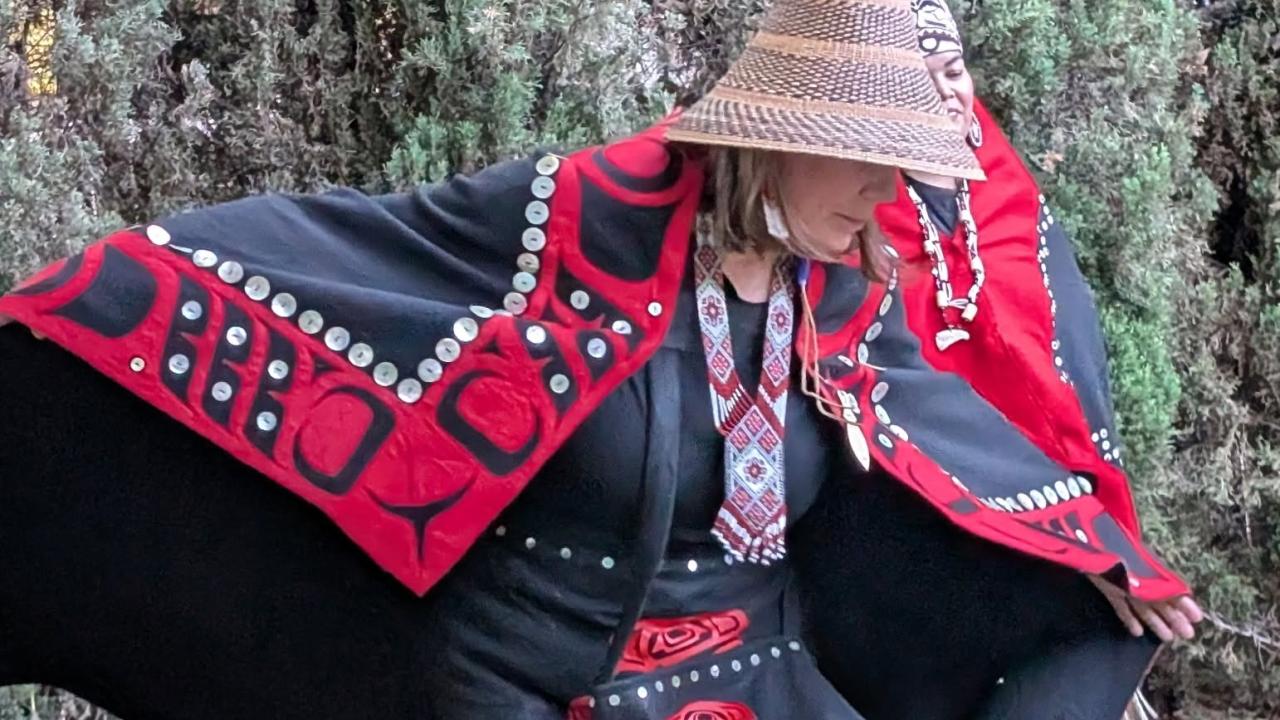 Person in red-and-black woven cloak and straw hat striking a low dance pose on grass