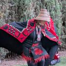 Person in red-and-black woven cloak and straw hat striking a low dance pose on grass